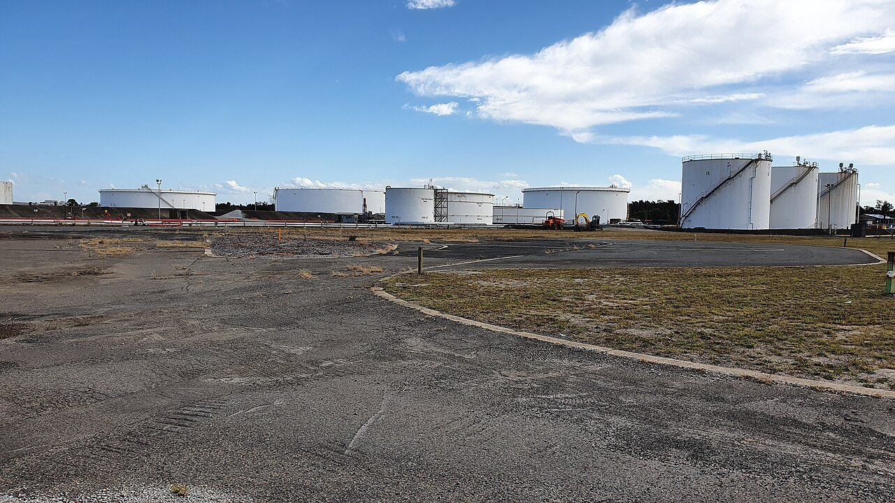 Battery of refinery storage tanks with associated pipework — storage and terminal assets carry continuous throughput exposure when shut down